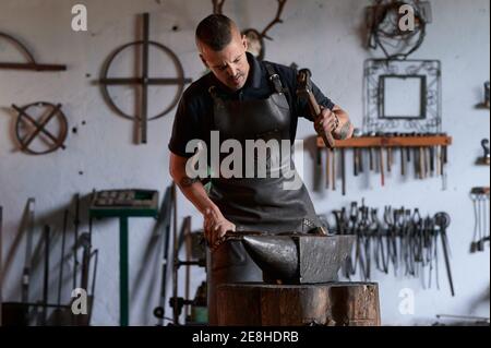 Concentrato giovane fabbro bearded in grembiule e occhiali di protezione colpisce metallo riscaldato con martello sull'incudine mentre si lavora in fucina Foto Stock