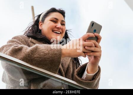Basso angolo di giovane allegra grumo femmina in chatting cappotto sul cellulare mentre si appoggia su recinzione in città Foto Stock