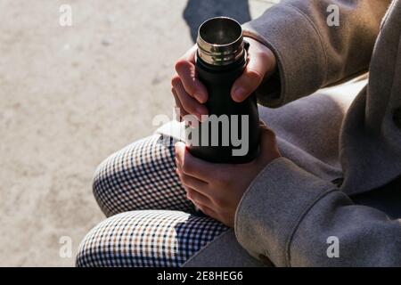 Femmina ad angolo alto che indossa abiti caldi che tengono aperto riutilizzabile bottiglia d'acqua sulle ginocchia mentre si siede sulla panca al sole via d'autunno Foto Stock