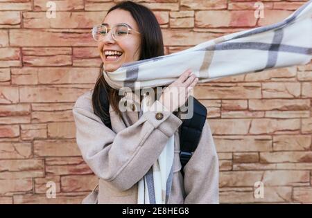 Eccitato giovane femmina in caldo cappotto e occhiali con zaino indossando una sciarpa volante accogliente mentre si è in piedi sulla strada passerella Foto Stock