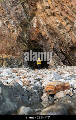 Vista posteriore anonimo viaggiatore in caldo giallino in piedi vicino ingresso alla grotta rocciosa situata su un terreno roccioso roccioso Foto Stock