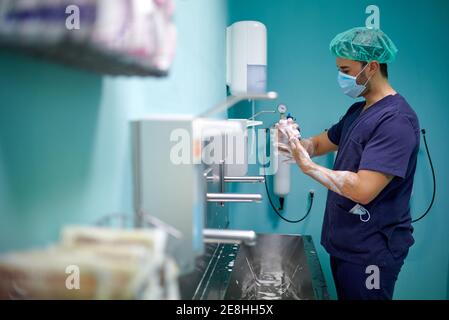 Vista laterale di anonimo giovane medico maschile in uniforme medica e maschera lavando le mani con sapone prima di un intervento chirurgico camera Foto Stock