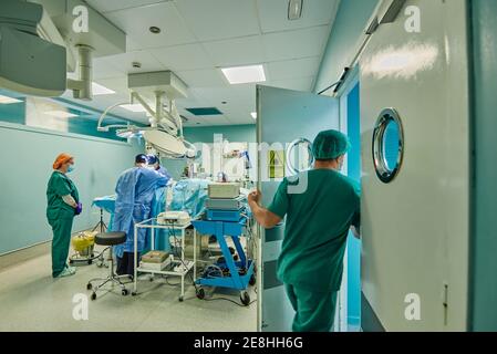 Vista posteriore di un assistente medico non riconoscibile che lascia la sala operatoria mentre gruppo di medici che eseguono interventi chirurgici in clinica moderna Foto Stock