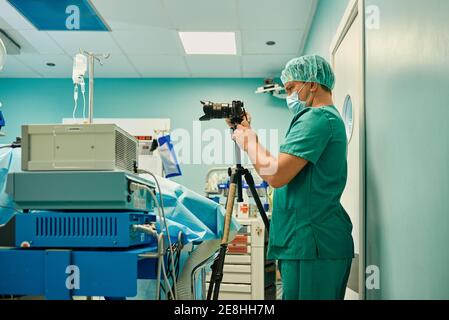 Vista laterale di un giovane fotografo medico maschile irriconoscibile in sterile maschera e uniforme per scattare foto con una fotocamera professionale durante l'intervento chirurgico in moder Foto Stock