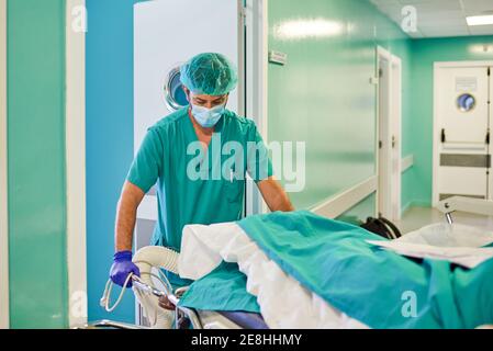 Giovane praticante maschile irriconoscibile in uniforme medica e maschera di trasporto paziente anonimo sdraiato su barella nel moderno corridoio ospedaliero Foto Stock