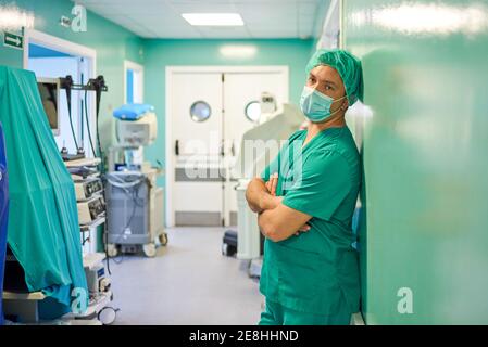 Vista laterale del medico giovane stanco in scrub e. maschera medica appoggiata a parete con braccia ripiegate mentre si riposa nel corridoio ospedaliero dopo l'intervento chirurgico Foto Stock