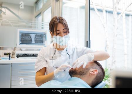 Dentista professionista concentrato in uniforme e maschera di controllo maschile pazienti i denti si sono condizionati durante il lavoro in clinica dentale moderna Foto Stock