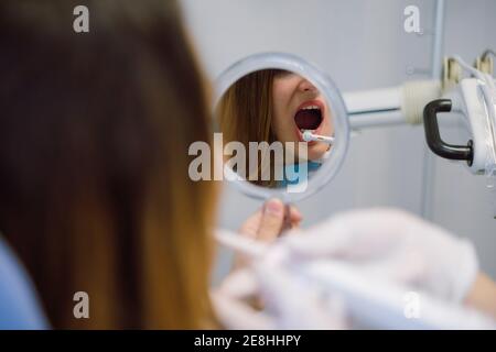 Vista posteriore raccolto giovane paziente femminile seduta su sedia dentale con la bocca aperta e guardando i denti nello specchio mentre dentista professionista in visita a mo Foto Stock