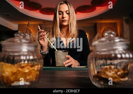 Biondo attraente barista femminile mescolando cocktail di alcol ghiacciato con lungo paglia che serve su bancone di legno Foto Stock