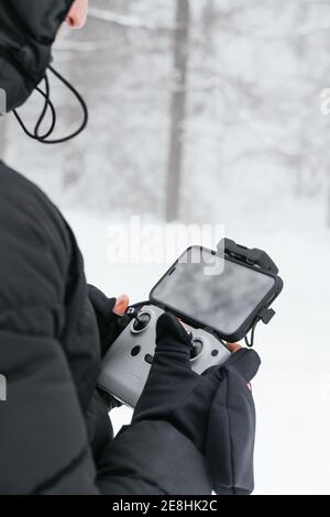 Giacca nera calda da uomo dal taglio non riconoscibile con cappuccio in piedi su terreni innevati con telecomando drone Foto Stock