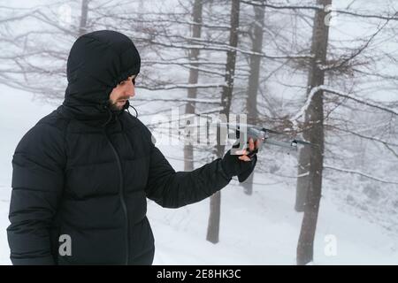 Concentrato giovane maschio in abiti caldi invernali e cappuccio di tenuta UAV contemporaneo in foresta ghiacciata nevosa Foto Stock