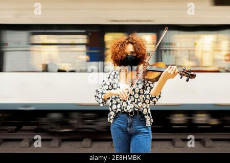 Attraente calmo musicista femminile in abbigliamento casual e viso protettivo mascherare il violino e guardare la fotocamera mentre si sta in piedi platfor stazione ferroviaria Foto Stock