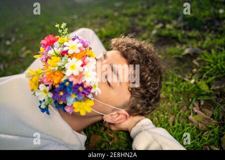 Dall'alto di anonimo giovane maschio con fiori colorati in fiore maschera sul viso e occhi chiusi sdraiati sul prato luce solare Foto Stock