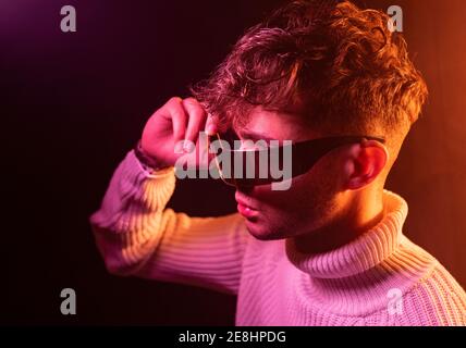 Vista laterale di un giovane maschio autoassicurato in un outfit elegante regolare gli occhiali da sole futuristici mentre ci si trova in uno studio buio con il neon illuminazione Foto Stock