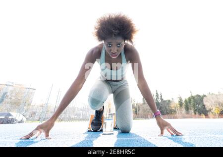 Da sotto di African American femmina corridore in blocchi di partenza in piedi in posizione di accovacciamento mentre si prepara per sprint a. stadio durante l'allenamento Foto Stock