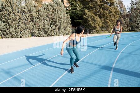 Allenati con le atlete multirazziali che passano il testimone mentre corri lungo la pista allo stadio durante la gara di relay Foto Stock