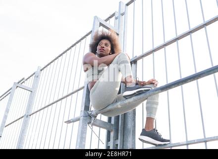 Da sotto di atleta afroamericana in abbigliamento sportivo seduto su barra orizzontale in metallo durante l'allenamento su terreni sportivi e. guardando via Foto Stock