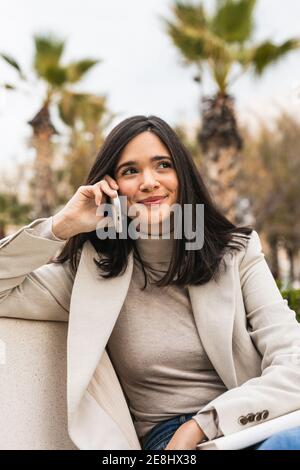 Allegra imprenditore femminile seduto su panchina in città e parlando sul telefono cellulare mentre sorridi e guarda via Foto Stock