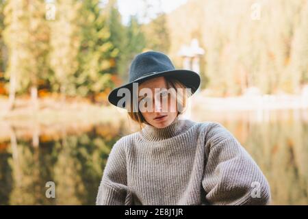 Vista laterale del corpo intero giovane positiva femmina in cappotto nero e cappello con zaino che cammina sul crosswalk in città alla luce del giorno Foto Stock