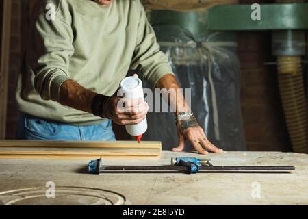 Falegname maschio maturo di raccolto irriconoscibile applicando colla su pezzo di legno mentre lavora a tavola in officina di falegnameria Foto Stock