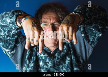 Allegro maschio di mezza età con capelli ricci e tatuaggi su mani senza un pollice in outfit trendy e occhiali sorridenti e mostrando movimento rock n roll Foto Stock