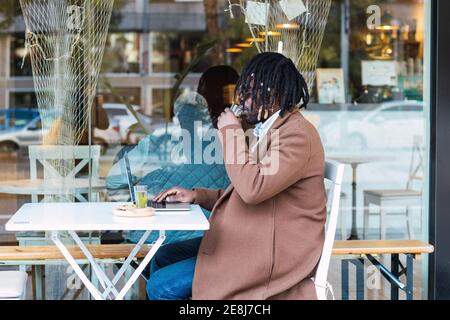 Vista laterale di elegante hipster afroamericano maschio con dreadlock lavorare in remoto con il computer portatile e bere bevande mentre si è seduti a. tavolo su restau Foto Stock