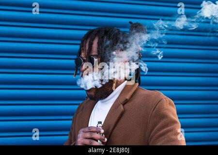 Moderno hipster adulto afroamericano maschio in abito alla moda e. in occhiali da sole con dreadlock fumare sigaretta elettronica contro parete blu Foto Stock