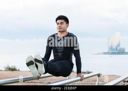 Corpo pieno di giovane atleta maschile asiatico in abbigliamento sportivo fare L Sit esercizio su barre parallele durante l'allenamento calistenico campo sportivo Foto Stock