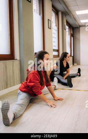 Sorridente allenatore femminile in abbigliamento sportivo che aiuta la ragazza allegra con stretching gambe mentre si riscaldano prima di allenarsi in forma fisica Foto Stock