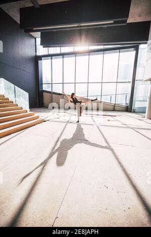 Vista laterale di un'artista teatrale femminile irriconoscibile che danzava con rialzato gamba e braccio sul pavimento con ombra contro parete di vetro Foto Stock