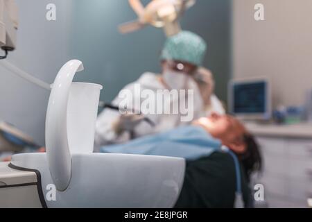 Bicchiere d'acqua nell'ufficio odontoiatrico. Medico femminile che si reca ad un paziente di sesso maschile. Mettere a fuoco la tazza di plastica bianca. Foto Stock