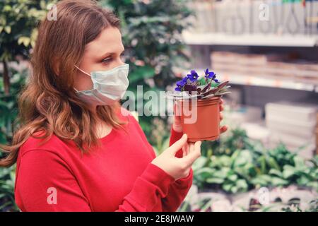 Donna in una maschera di faccia a una serra con fiori viola blu in una pentola. Vendite in negozio fiori in vaso per un hobby casa durante la pandemia coronavirus Foto Stock