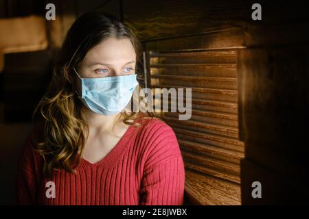 Una donna in una maschera viso si trova in una finestra leggermente aperta e si affaccia, un ritratto durante il periodo di quarantena Foto Stock