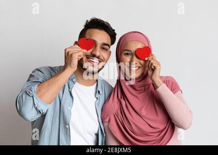 Concetto di San Valentino. Coppia musulmana amorevole che tiene i cuori di carta rossa vicino agli occhi Foto Stock