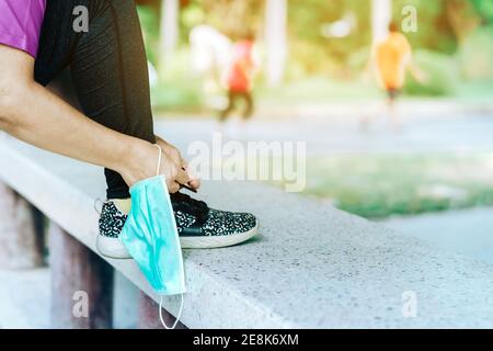 Fitness donna togliere la sua maschera chirurgica prepararsi a fare esercizio di allacciatura scarpe da corsa per jogging all'aperto. Nuovo stile di vita normale. Assistenza sanitaria Foto Stock