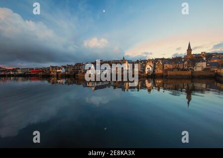 Vista grandangolare della cittadina di pescatori Stromness sulle isole Orkney con la luna che riflette in acqua e edifici in pietra distandt a. alba Foto Stock