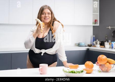 Concetto di dieta e nutrizione. Giovane donna dal corpo curvo felice con lunghi capelli biondi usando il frullatore nella cucina moderna, mescolando la frutta fresca per sano Foto Stock