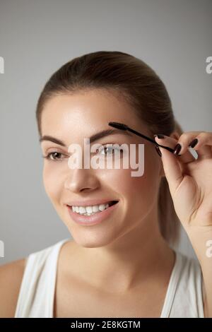 Trucco di bellezza. Bella donna che forma sopracciglia closeup. Modello della ragazza con trucco professionale usando spazzola di sopracciglia e sorridente Foto Stock