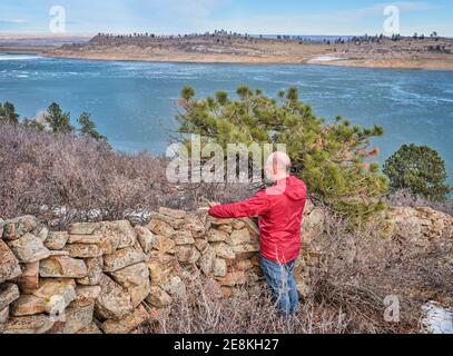 escursionista senior presso la vecchia recinzione di pietra che si affaccia sul lago di montagna congelato - serbatoio Horsetooth ai piedi delle Montagne Rocciose - A. popolare area ricreativa Foto Stock