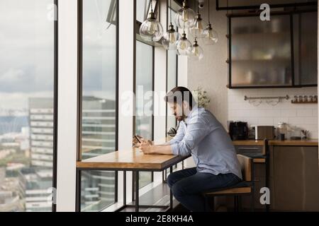 Un giovane sorridente che si rilassa vicino a una grande finestra panoramica usando uno smartphone Foto Stock