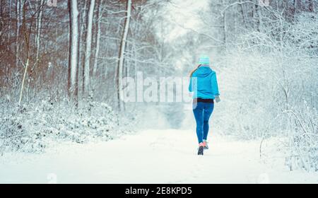 Giovane donna che corre lungo un sentiero nei boschi invernali attraverso la neve Foto Stock