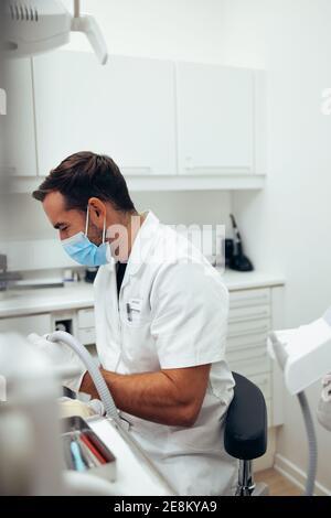 Dentista maschile che tratta un paziente in odontoiatria. Medico dentale che indossa la maschera di faccia seduto nella sua sedia presso la clinica dentale. Foto Stock