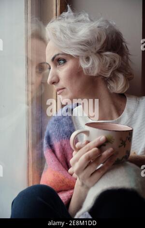 Infelice donna matura che beve tè e guardando fuori finestra, blocco e concetto di isolamento, fuoco selettivo Foto Stock