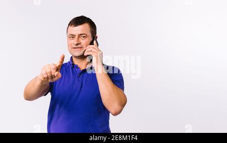 L'uomo anziano parla sullo smartphone e punta il dito indice verso la fotocamera. Sfondo grigio e spazio pubblicitario vuoto. Foto Stock