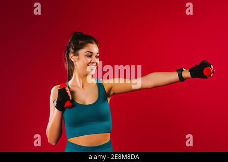 Ragazza bruna caucasica sportiva con manubri e un sorriso si esercita su uno sfondo rosso e guarda al lato. Foto Stock