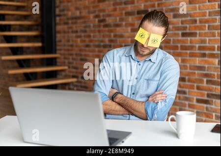 Un giovane lavoratore d'ufficio stanco in una camicia casual seduto alla scrivania sul suo posto di lavoro, mettere gli adesivi sugli occhi per nascondere che sta annidando o. Foto Stock