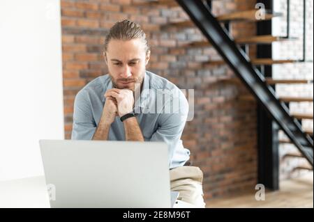 Pensieroso giovane imprenditore maschile che lavora sul progetto online sul portatile, ha messo le mani sul mento, guardando lo schermo, sentendosi sorpreso Foto Stock