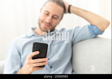 Attraente giovane hipster sopportato millenario che si rilassa e si siede sul comodo divano nel soggiorno, chiacchierando con un amico sui social media Foto Stock