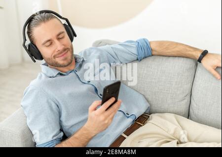 Attraente giovane hipster sopportato millenario indossando le cuffie, distendersi e distendersi sul divano comodo, ascoltare la musica, chattare con Foto Stock