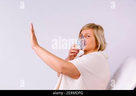 La donna caucasica senior inalare con una maschera nebulizzatrice a casa per la malattia di coronavirus, mostrando il segno di stop su sfondo grigio. Foto Stock